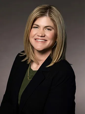 Professional portrait of a smiling woman with blonde hair, wearing a black blazer and green top.