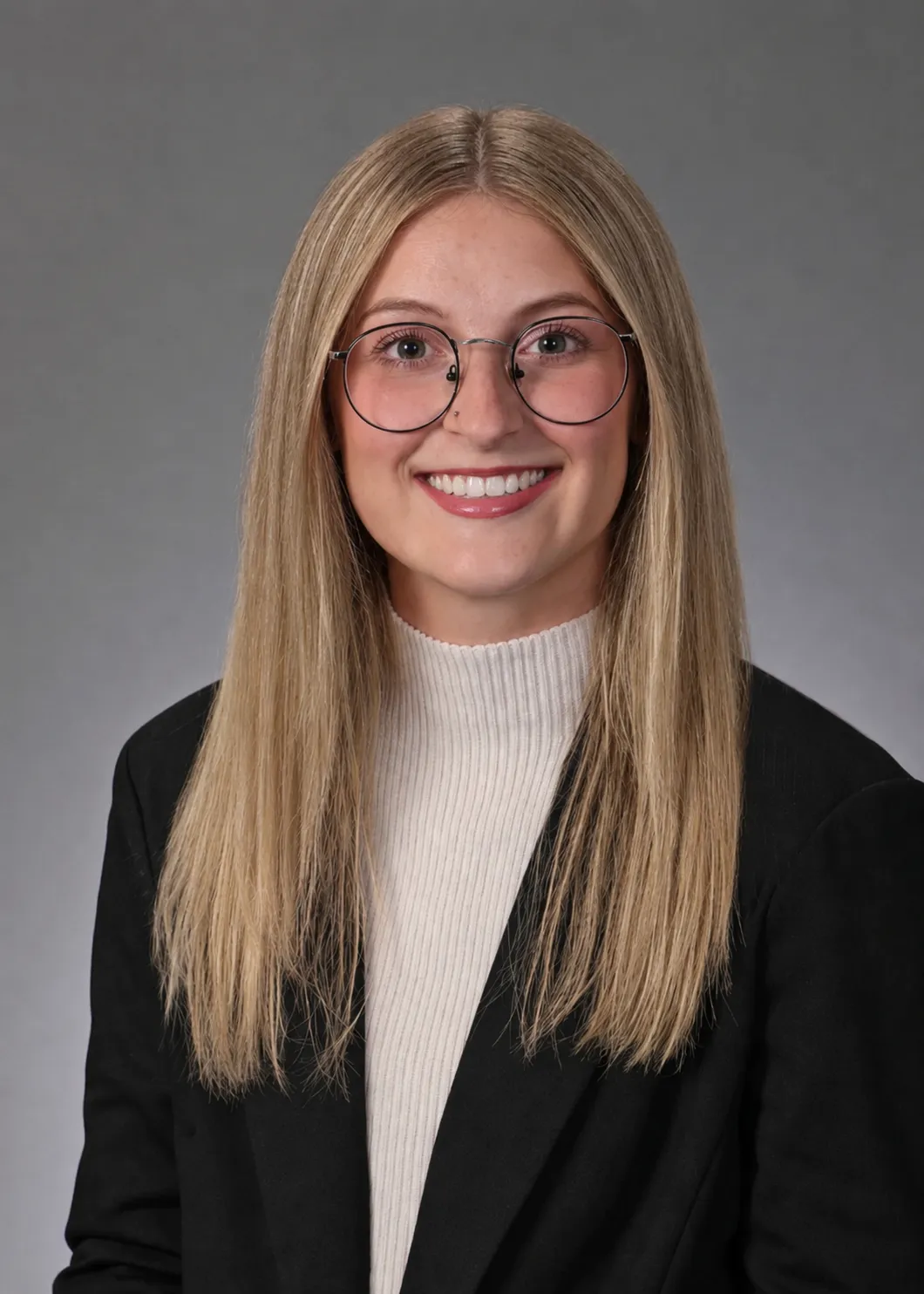 Professional headshot of a smiling young woman with long blonde hair and round glasses, wearing a black blazer and white turtleneck.