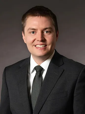 Professional headshot of a smiling man in a dark suit, white shirt, and gray tie.