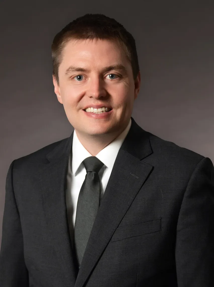 Professional headshot of a smiling man wearing a dark suit, white shirt, and black tie against a neutral background.