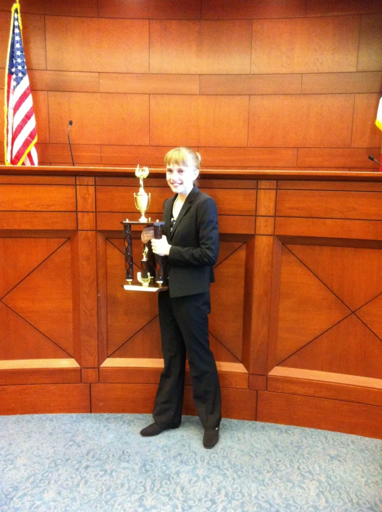 Young girl in formal suit holding large trophy in courtroom setting with American flag
