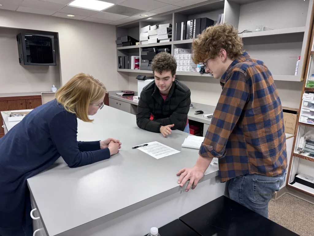 Three people engaged in discussion around a gray office table with documents in a storage room.