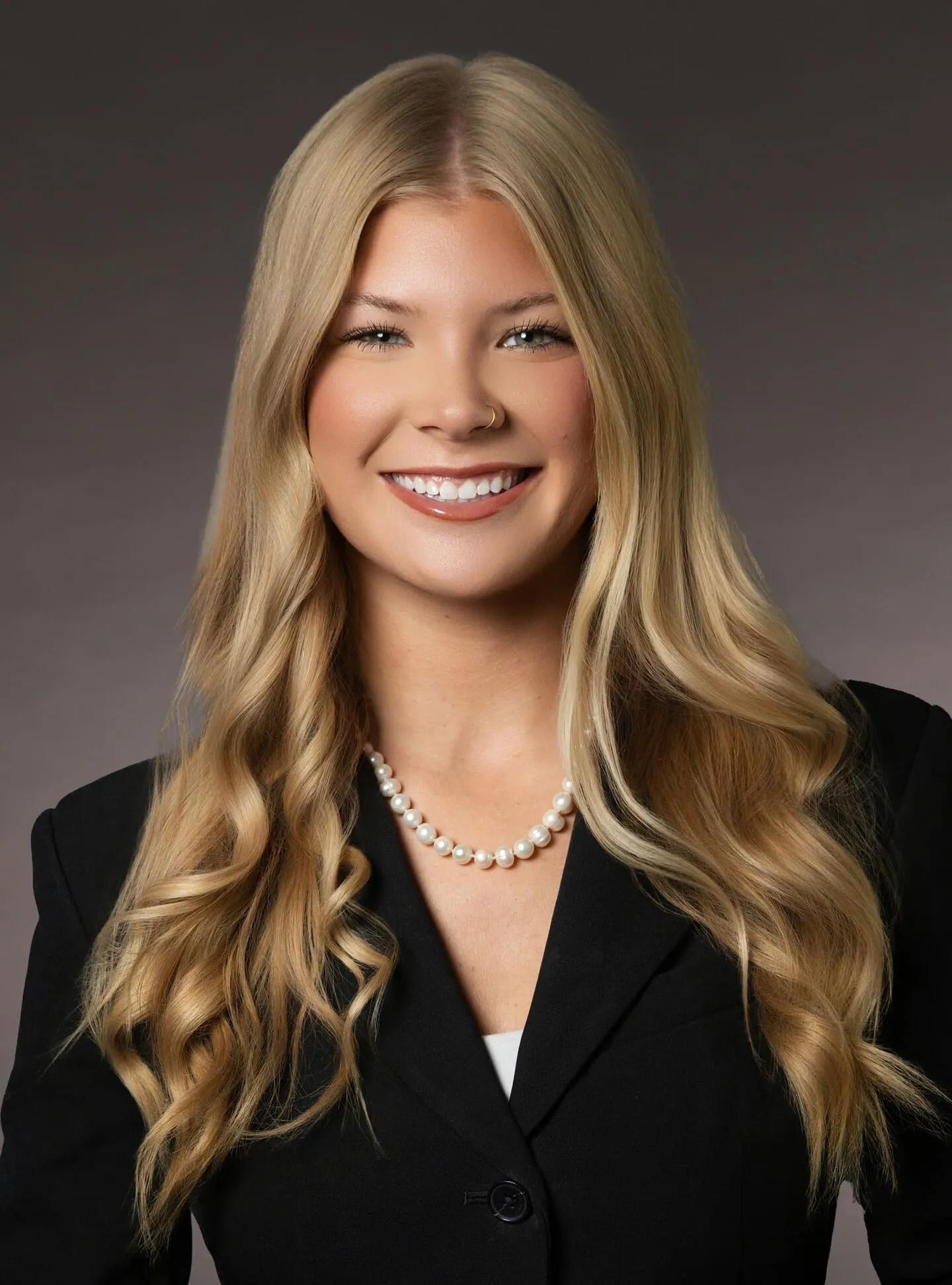 Professional portrait of a smiling blonde woman with long wavy hair, wearing a black blazer and pearl necklace.